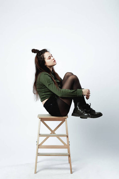Stylish young dark-haired girl with a funny hairstyle dressed in green shirt, black shorts , tights and shoes is posing sitting on the wooden stool on the white background in the studio - Photo, image