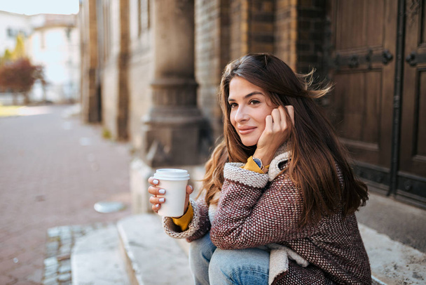 Brunette sitting on stairs holding a take away coffee cup. - 写真・画像