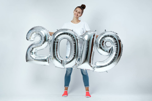 Happy young girl dressed in white t-shirt, jeans and pink socks holding balloons in the shape of numbers 2019 on the white background in the studio - Fotó, kép
