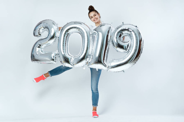 Happy young girl dressed in white t-shirt, jeans and pink socks holding balloons in the shape of numbers 2019 on the white background in the studio - Fotografie, Obrázek
