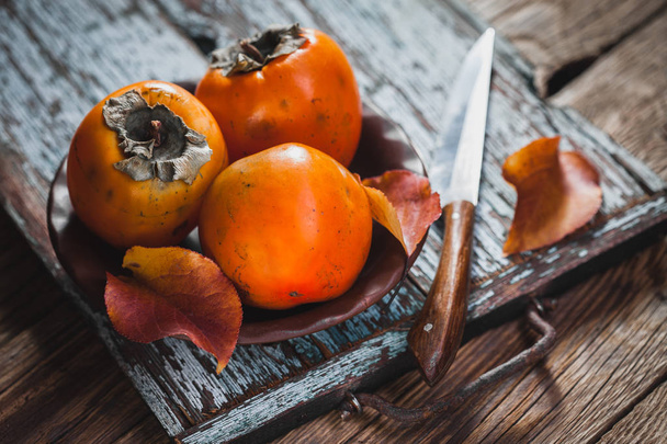 Ripe orange persimmon fruit and persimmon leaves in a brown plate on a brown wooden table. fresh fruits - Foto, Imagen