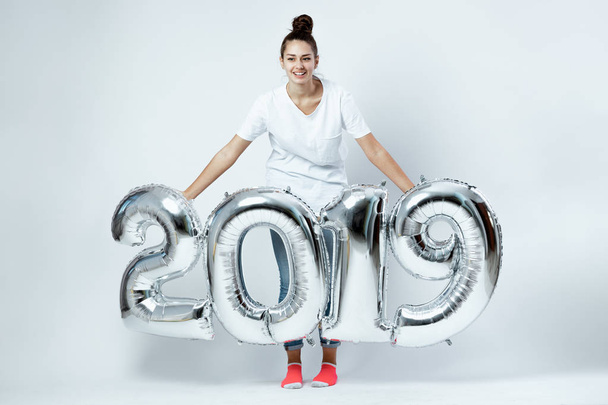 Smiling young girl dressed in white t-shirt, jeans and pink socks holding balloons in the shape of numbers 2019 on the white background in the studio - Photo, Image