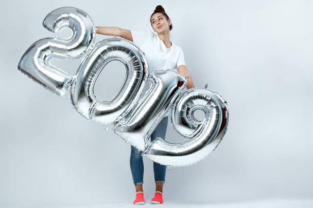 Funny young girl dressed in white t-shirt, jeans and pink socks holding balloons in the shape of numbers 2019 on the white background in the studio - 写真・画像