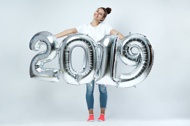 Smiling young girl dressed in white t-shirt, jeans and pink socks holding balloons in the shape of numbers 2019 on the white background in the studio - Fotó, kép