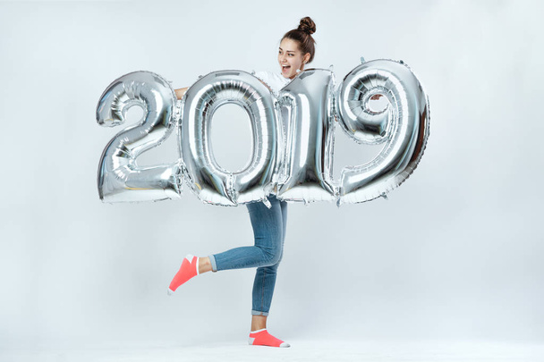 Funny young girl dressed in white t-shirt, jeans and pink socks holding balloons in the shape of numbers 2019 on the white background in the studio - Photo, Image