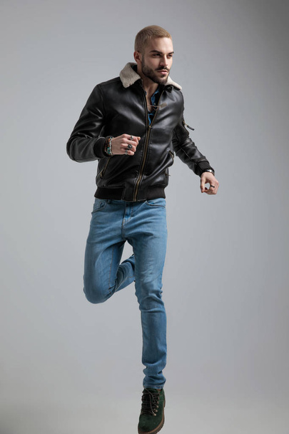Mann in Lederjacke und Jeans springt und schaut vor grauem Hintergrund weg - Foto, Bild