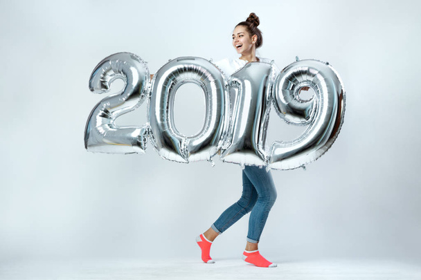 Young funny girl dressed in white t-shirt, jeans and pink socks holding balloons in the shape of numbers 2019 on the white background in the studio - Photo, Image
