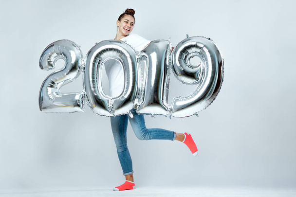 Young funny girl dressed in white t-shirt, jeans and pink socks holding balloons in the shape of numbers 2019 on the white background in the studio - Photo, Image