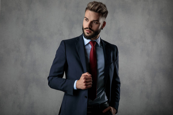 relaxed businessman holding suit's collar and hand in pocket looks to side while posing in studio - Foto, afbeelding