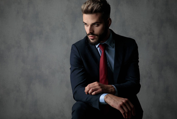 giovane uomo d'affari attraente appoggiato gomito sul ginocchio guarda a lato su sfondo grigio
 - Foto, immagini