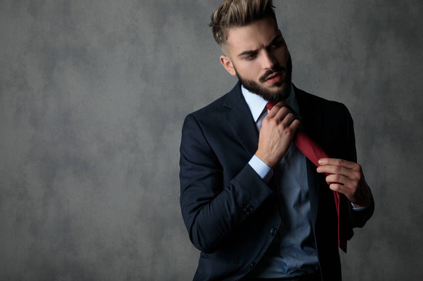 seriöser Geschäftsmann zieht Krawatte und blickt zur Seite auf grauem Hintergrund - Foto, Bild