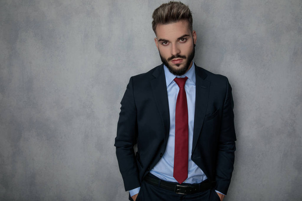 relaxed young fashion businessman standing with hands in pockets against grey studio wal - Photo, Image