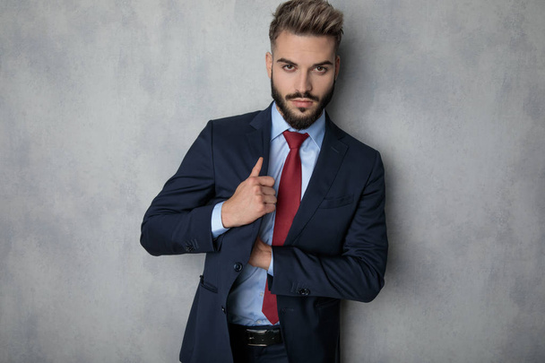 jovem empresário sexy procurando no bolso interior de seu casaco, enquanto está de pé contra a parede cinza
 - Foto, Imagem
