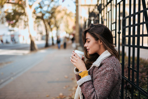 Young stylish woman drinking hot drink in autumn city street. - 写真・画像