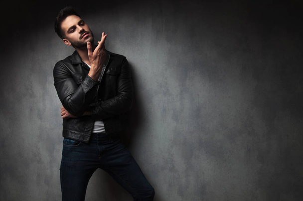 arrogant sexy young man in leather jacket is gesturing to a side while posing in studio - Φωτογραφία, εικόνα