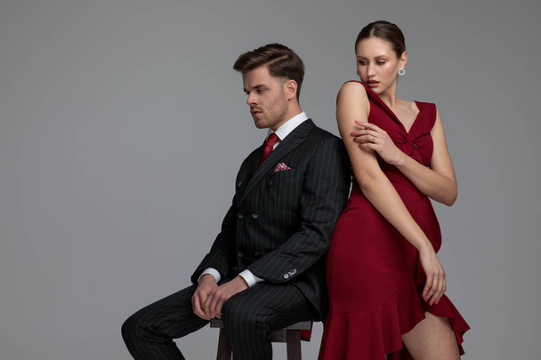 elegantní pár zády k sobě, pózuje v studio, muž sedí na židli a žena dotýkat rukama na šedém pozadí - Fotografie, Obrázek