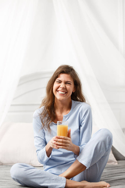 Happy brunette girl dressed in the light-blue pajama sits on the bed with glass of fresh juice in her hands and smiles in the cozy light room - Foto, imagen