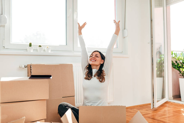 Moving to new home. Happy woman with arms raised sitting on the floor among carton boxes. - Foto, afbeelding