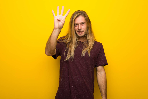 Блондин с длинными волосами над желтой стеной счастлив и считает четверых с пальцами
 - Фото, изображение