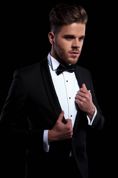 young elegant man puliing his tuxedo's collars while looking away from the camera on black background - Foto, afbeelding