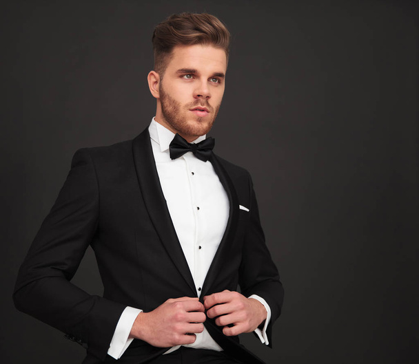 side view of young groom buttoning his coat and looking away on black background - Photo, Image