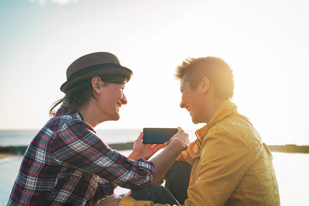 Joyeux couple lesbien regardant sur le téléphone mobile à côté de la plage au coucher du soleil - Les jeunes femmes homosexuelles s'amusent avec les nouvelles technologies tendances - Lgbt, homosexualité, concept de technologie et de relation
 - Photo, image