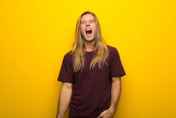Blond man with long hair over yellow wall shouting to the front with mouth wide open - Foto, afbeelding