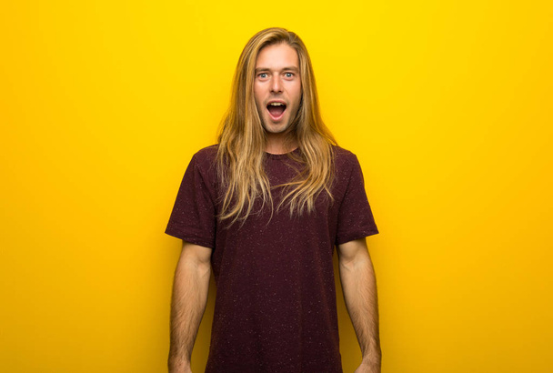 Hombre rubio con el pelo largo sobre la pared amarilla con sorpresa y expresión facial impactada
 - Foto, Imagen