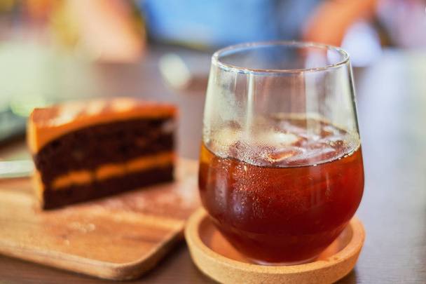 Eisschwarzer Kaffee im Glas im Vordergrund und weiche Unschärfe für Schokolade-Karamell-Käsekuchen auf Holzteller - Foto, Bild