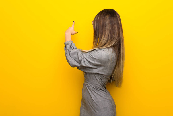 Молода жінка в окулярах над жовтою стіною вказує назад з індексним пальцем
 - Фото, зображення