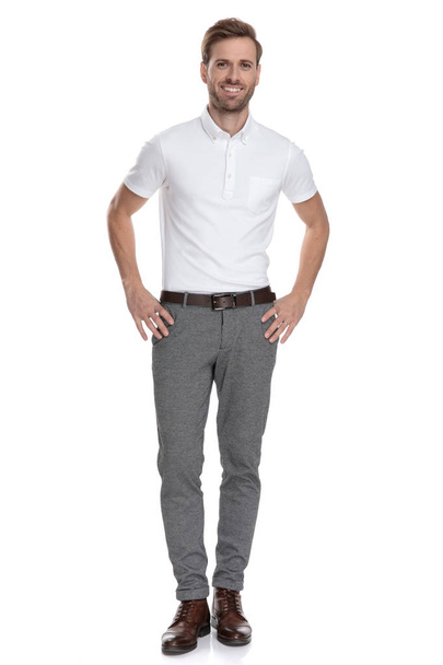 Happy smart casual jongeman met handen op taille staande op witte achtergrond - Foto, afbeelding