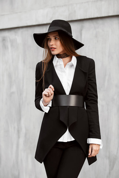 Fashionable slim long-haired girl dressed in a black stylish jacket on a white shirt, black pants and a hat with a wide brim poses in the street - Foto, afbeelding