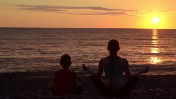 Äiti ja poika meditoivat rannalla lootusasennossa. Näkymä takaa, auringonlasku, siluetit
 - Materiaali, video