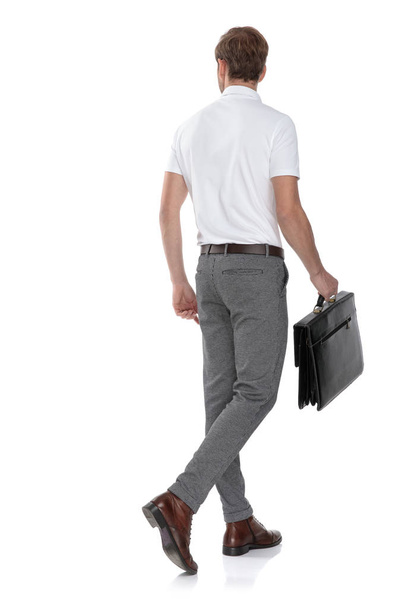 Achteraanzicht van een smart casual jongeman wandelen met een werkmap en kijkt naar de kant op witte achtergrond - Foto, afbeelding