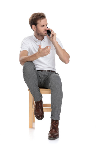 mylić mężczyzna siedzi i gestykulacji w zamieszanie podczas rozmowy na jego telefon na białym tle - Zdjęcie, obraz