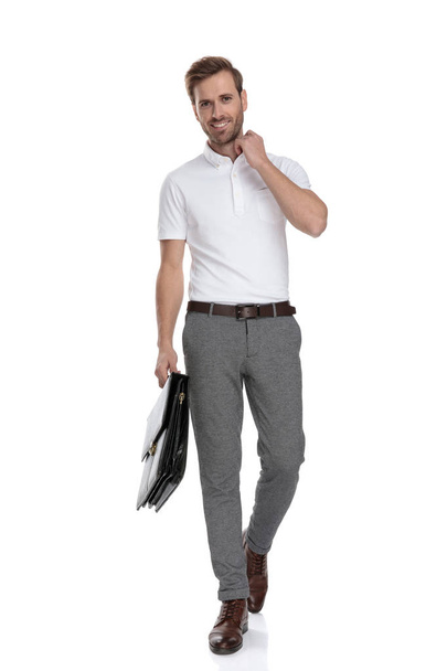 sorridente giovane uomo casual sta camminando con valigetta e regola il suo colletto su sfondo bianco
 - Foto, immagini