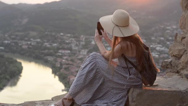 Meisje in een hoed met een rugzak zittend op de heuvel. Schiet video naar smartphone, stad hieronder - Video