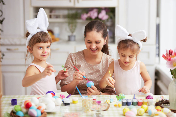 Happy young mother and her two little daughters with white rabbits ears on their heads dye the eggs for the Easter table in cozy light kitchen - Фото, изображение