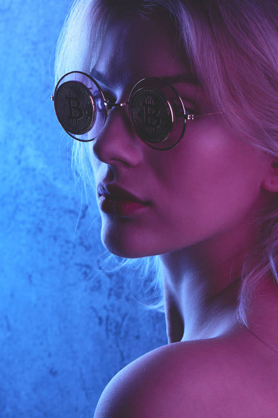 Perfil de una hermosa joven rubia con labios rojos en gafas con bitcoins en tono rojo azul. Concepto criptomoneda. Iluminación creativa de neón
 - Foto, Imagen