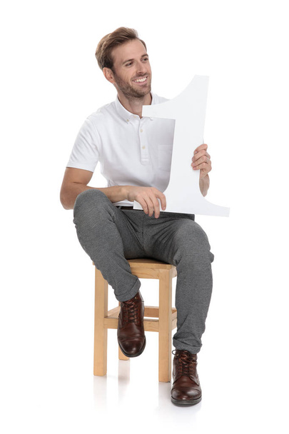 lächelnd sitzt ein lässiger Mann mit einem Buchstaben mit der Nummer eins und blickt zur Seite auf weißem Hintergrund - Foto, Bild