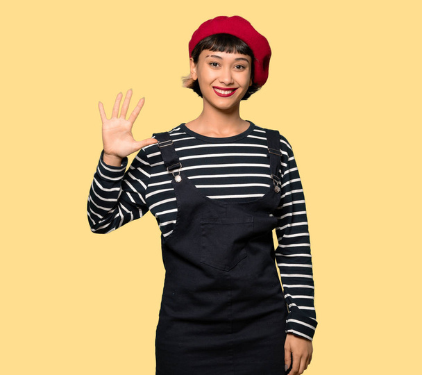 Young woman with beret counting five with fingers over yellow background - Фото, изображение