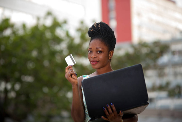 Smiling and confident businesswoman doing online shopping through a laptop and a credit card standing in the street - Photo, Image