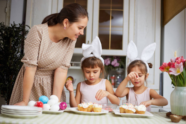 Young mother and her two little daughters with white rabbits ears on their heads cook small Easter cakes for the Easter table in the cozy light kitchen - Фото, изображение