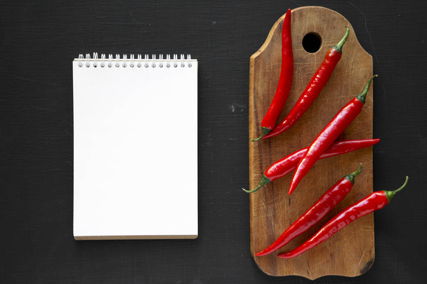 Hot red chili peppers and blank notepad on black background, overhead view. Space for text. - Photo, Image