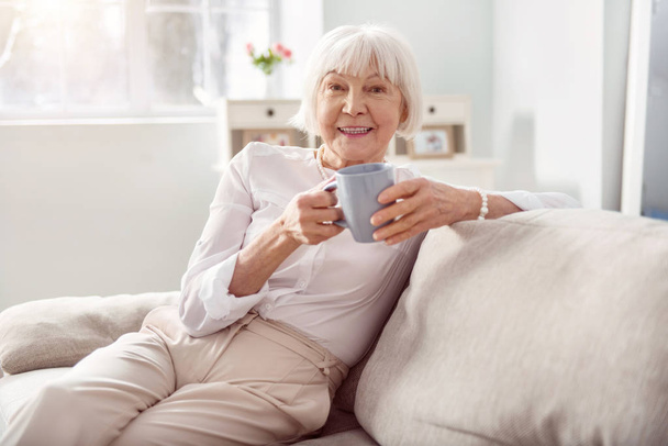 Upbeat senior woman drinking coffee on sofa - Valokuva, kuva