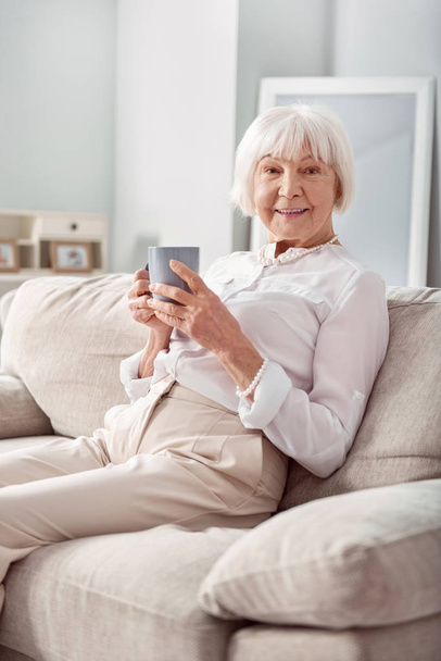Charming senior woman posing with cup of tea on couch - Foto, Bild