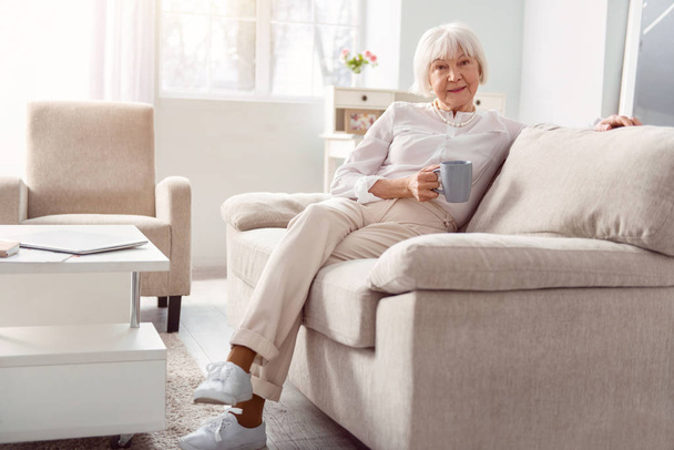 Charming elderly lady relaxing with coffee cup on couch - Photo, image