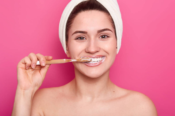 Dental care concept. Pleased young woman has brilliant smile white shining teeth, cleans teeth with wooden toothbrush and whitening toothpaste, isolated over pink background, has natural beauty. - Foto, Imagem