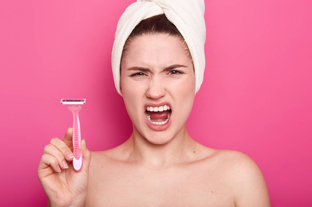 Displeased angry woman against of shaving, holds razor in hand, supports wax depilation, poses half nude, shows bare shoulders, frowns face, isolated over pink background. Women and hygiene concept - Foto, Imagem