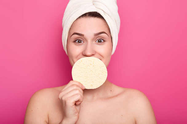 Horizontal shot of pleased woman with satisfied expression, covers mouth with sponge, looks at camera in satisfaction, cares of body and skin, has white towel on head, isolated over pink background - Photo, image